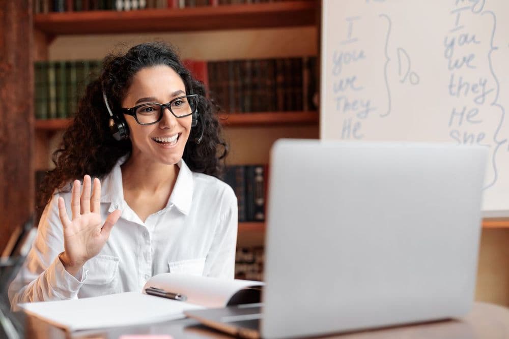 Professora de inglês online com headset, sorrindo durante uma aula virtual, em um ambiente de estudos com livros e quadro branco ao fundo, ensinando verbos em inglês.