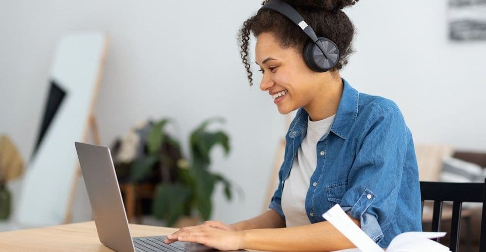 Mulher jovem sorridente usando fones de ouvido enquanto descobre no notebook como saber seu nível de inglês.