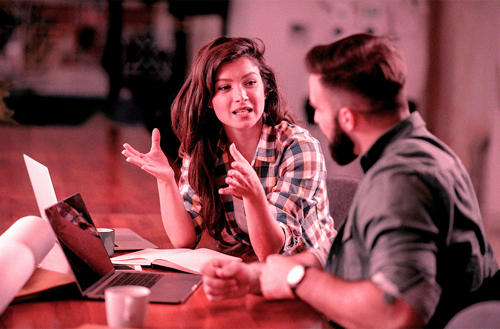 Mulher conversa com homem durante uma reunião em escritório, ambos sentados à mesa com laptop e documentos, praticando estruturas do simple past em uma atividade de inglês profissional.